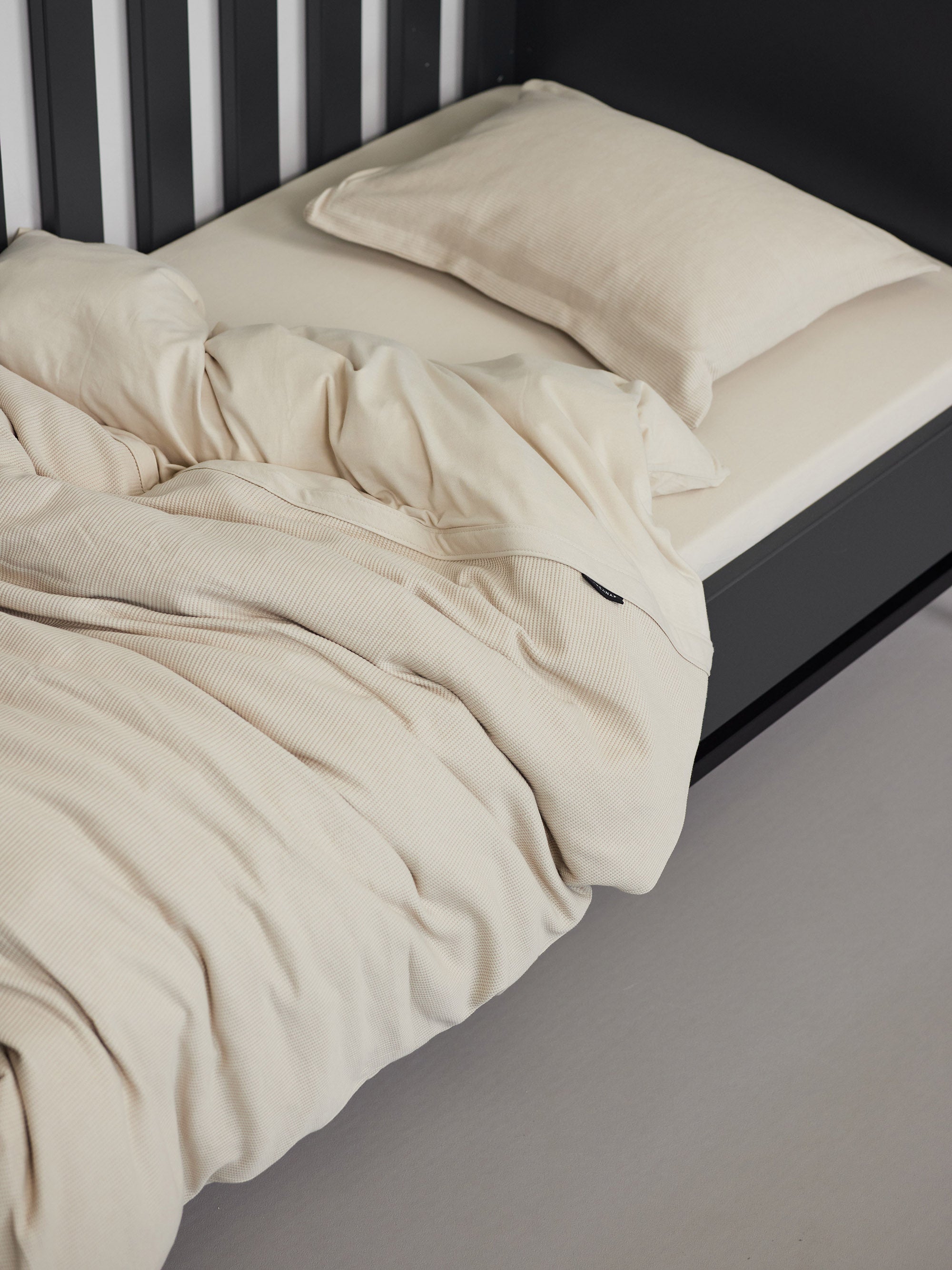 Hey welcome 2 JACQUARD | Sand Toddler Pillowcase. Image: Duvet, pillow and pillowcase on a black crib with sand sheet